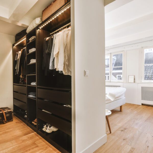 Modern spacious walk-in closet with dark wood shelving, warm LED lighting, and an open-concept transition to a bright, Scandinavian-style bedroom.