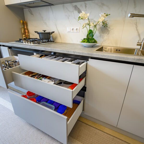 A sleek modern kitchen with white and gray cabinets, featuring open deep storage drawers neatly organized with kitchen utensils and containers.