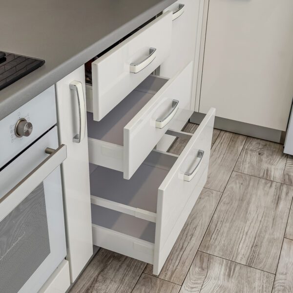 A modern white kitchen with an open drawer system, featuring sleek handles, a built-in oven, and wood-look flooring for a stylish and functional design.